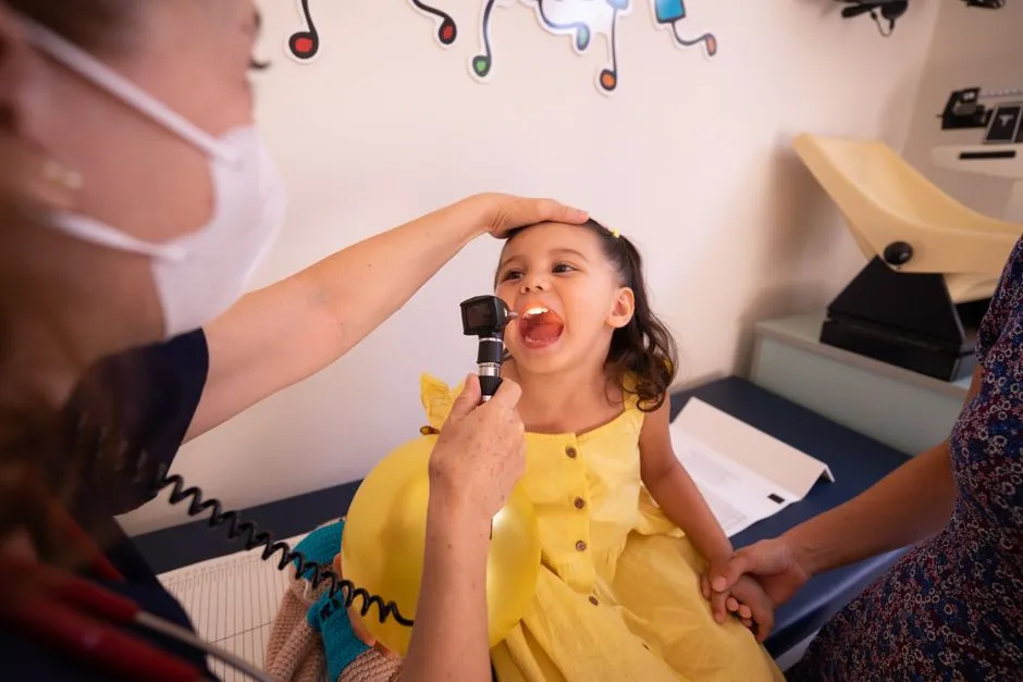 A pediatrician examining a young child's throat during a checkup, parent standing nearby